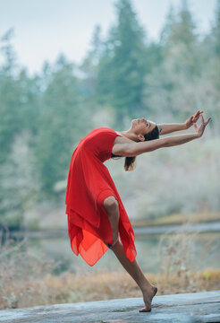 Woman in a red dress dancing gracefully outdoors with a forest background. USA