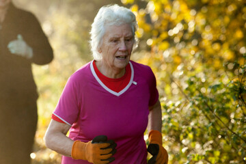 An elderly woman with white hair jogs energetically in a park, wearing a bright pink shirt and orange gloves. Sale Water Park, Manchester, UK