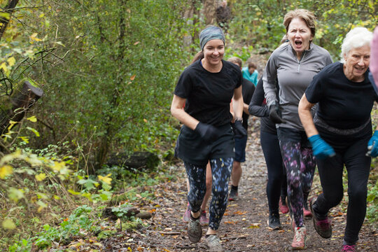 A group of women of varying ages are energetically running through a forest trail in autumn, surrounded by green and yellow foliage. Sale Water Park, Manchester, UK