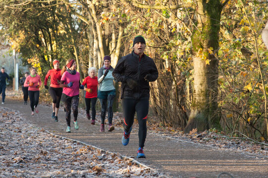 A group of people jogging on a path covered with fallen leaves in a forest, wearing colorful athletic clothing. Sale Water Park, Manchester, UK