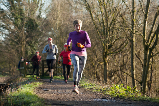 A group of runners jog along a sunny forest trail dressed in winter athletic wear, with leafless trees lining the path. Sale Water Park, Manchester, UK