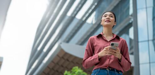 Successful asian business woman using phone outdoors.Happy businesswoman holding cellphone using smartphone or mobile phone standing or walking on city urban street,6g communicating with a colleague.

