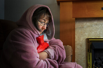 Obraz premium Senior Woman Wrapped in Dressing Gown in Unlit and Unheated Home Holding Hot Water Bottle During Winter Heating Crisis