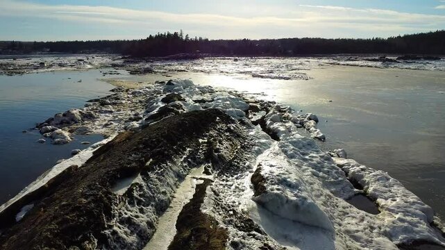 Backlit drone view of a rocky shoreline in Mitis Bay with sunlight reflecting across frozen surfaces toward the dark forest edge in Metis‑sur‑Mer, Quebec, Canada
