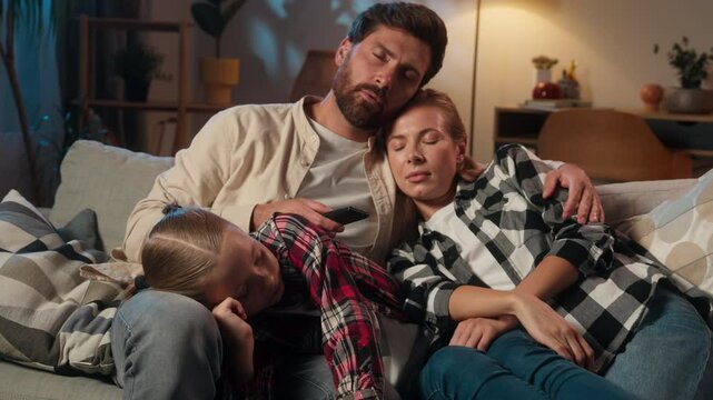 Tired sleepy exhausted fatigued Caucasian family mother woman man father with child kid girl daughter together sleeping napping relaxing on couch at home in evening dark night living room cozy sofa