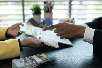 Two people are sitting at a table with a piece of paper in front of them. One of them is pointing at the paper. There are two calculators on the table, one on the left and one on the right
