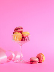 Pink and yellow color macarons in a champagne glass and pink ribbon on a pink background. Valentine's day card with a copy space