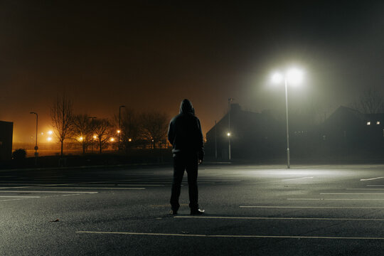 A sinister hooded figure, standing in a city car park. On a mysterious foggy night. Silhouetted by street lights