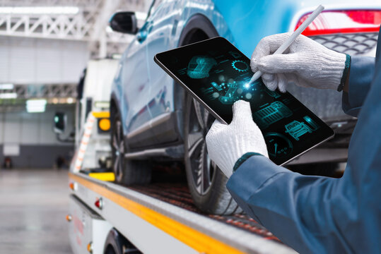 A mechanic wearing gloves uses a digital tablet with a futuristic interface to diagnose a car loaded onto a tow truck in a garage.