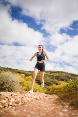 Female trail runner wearing black sportswear and a hydration vest, running through a scenic natural landscape on a rocky path.