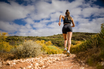 Female trail runner wearing black sportswear and a hydration vest, running through a scenic natural landscape on a rocky path.