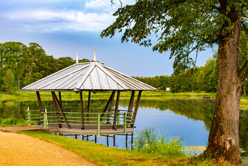  A wooden gazebo with a metal roof overlooking a calm lake surrounded by lush greenery and trees on a bright, peaceful day...