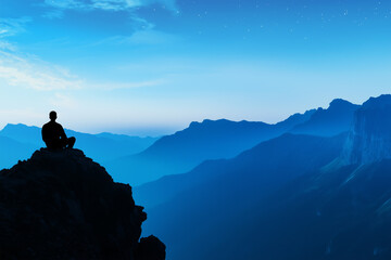 Person meditating on a rocky outcrop overlooking a serene mountain range at dusk