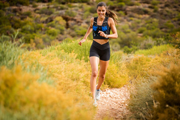 Female trail runner wearing black sportswear and a hydration vest, running through a scenic natural landscape on a rocky path.
