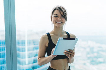 Fototapeta premium Half length portrait of beautiful slim woman in activewear holding modern digital tablet for blogging, cheerful hipster girl sending mails and communicating via tablet connected to 4G internet
