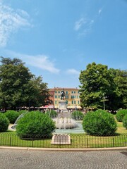 Obraz premium Beautiful historic town square in Europe featuring a water fountain, lush green topiary bushes, and an equestrian monument under a bright blue sky