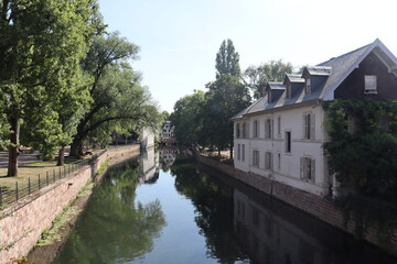 Fototapeta premium Peaceful riverside view in Strasbourg with historic house, calm canal waters, and lush green trees creating a serene summer atmosphere.