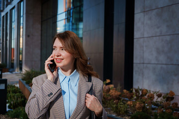 A cheerful young businesswoman in a stylish blazer is having a phone conversation outdoors in an urban setting. She is smiling confidently, exuding positivity and professionalism. 