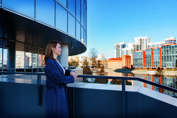 A young woman in a blue coat stands on a terrace, gazing at the modern city skyline with a calm and thoughtful expression. The glass building behind her reflects the urban landscape.