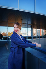 A stylish young woman in a blue coat stands outside a modern glass building, leaning on a railing and smiling. The reflective windows of the structure capture the surrounding cityscape.