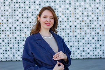 Elegant woman in a stylish blue coat standing against a modern architectural background. She smiles confidently, exuding warmth and sophistication. Outdoor portrait with natural lighting.