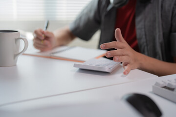 Business professional working at a desk, using a calculator and taking notes. A coffee mug sits nearby, suggesting a focused work environment in an office setting
