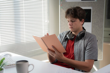Young man with headphones around his neck reading a document at a desk. Sunlight streams through...