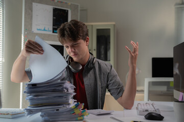 Young man in an office setting, overwhelmed by a large stack of paperwork. He looks frustrated,...