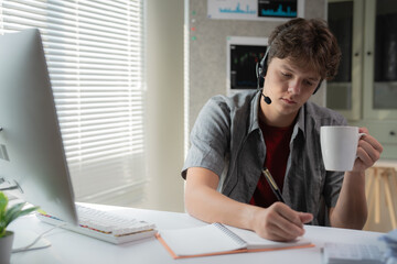 Young man wearing a headset, writing notes while holding a coffee mug, working at a desk with a computer. Bright natural light filters through the window blinds, creating a productive atmosphere