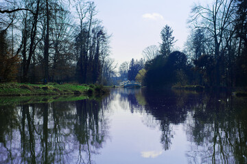 A serene river scene with reflections of bare trees on the water, a grassy riverbank, and a distant white bridge under a clear sky.