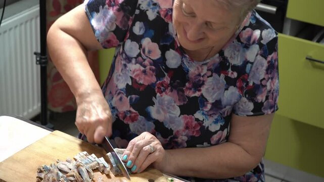 An elderly woman cuts herring fish with a knife, prepares food. at home in the kitchen. Old hands and knife close up. Wooden board.