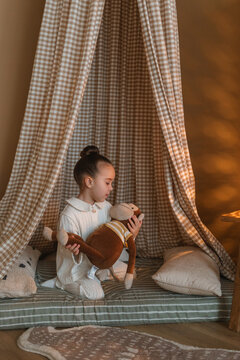 Young girl in white leisurewear sitting in a teepee in her bedroom playing with a toy stuffed monkey