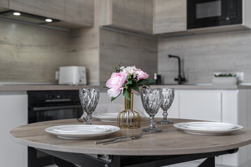 stylish modern kitchen design, wooden cabinets. dining table with table setting. white plates, glasses and flowers in vase on wooden table. Lunch at home. Modern built-in kitchen in minimalist style.
