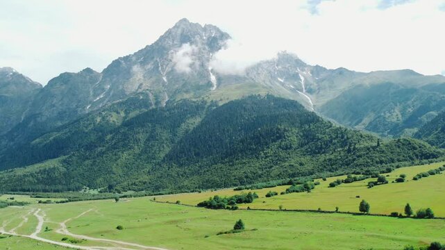 White clouds gently caress towering mountain peak, casting shadows on bright green valley below, where dirt road winds through picturesque landscape. Shooting from drone.