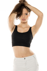 Fototapeta premium Dynamic fashion portrait showcasing model in black crop top and white pants with decorative details. Natural, energetic pose with hands styling hair against clean background