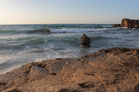Beach of luxury hotel. sunny day, windy, stormy, wavy, large rocks in sea, blue sky. shines soft yellow-gold brown-golden sand. Sea waves crashing on the shore with wavy white foam. island of cyprus