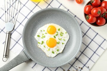 Tasty fried eggs with green onion in frying pan and tomatoes on white wooden table, flat lay