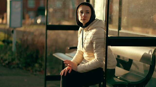 Woman waiting bus at public transport stop in quiet residential area. Female sitting on bench under shelter while looking down street. Lady resting outdoors at suburban bus stop during sunny day