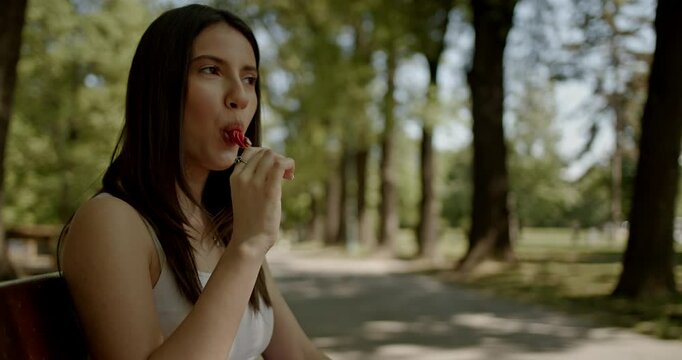 Young pretty woman sitting on a park bench enjoying a lollipop on a summer day. She's happy and smiling. 4K stock video great for advertisements and campaigns. - Powered by Adobe