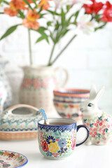 Ceramic mug with tea on the kitchen table with a jug with red flowers. Selective focus on the mug.