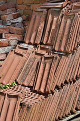 A pile of weathered, terracotta roof tiles are stacked against a brick wall, showcasing their rustic texture and earthy colors