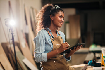 African American woman carpenter using a digital tablet in a wood factory workshop, planning designs and managing production workflow for a small business in the woodworking industry
