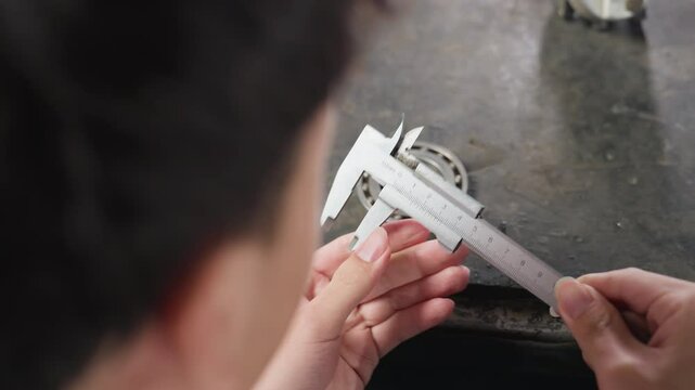 Overhead view of lab technician using vernier caliper to measure diameter of boris on workbench under dim light, recording measurements on tablet nearby. Industrial workspace with precision tools