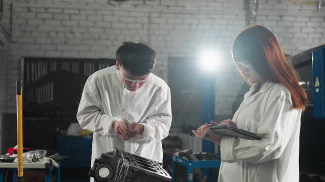 Two inventors in Chinese automotive workshop, boy showing reading to girl while she records data on tablet, he measures engine component again under bright light illuminating scene