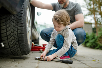 Cute little boy helping his father to change car wheels at their backyard. Father teaching his...