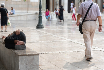 Homeless person sleeping on bench while tourist is walking away in city square