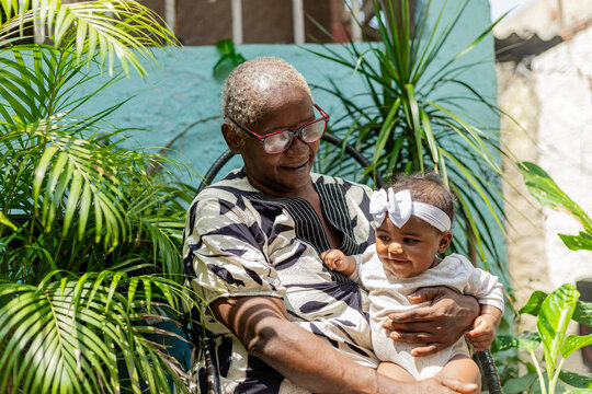 Happy black grandmotherr with her newborn granddaughter on her lap in a natural environment with plants. Rio de Janeiro, Brazil.