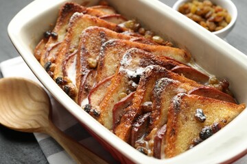 Freshly baked bread pudding in baking dish and wooden spoon on dark table, closeup