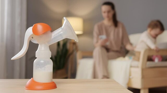 A mother multitasks by using a breast pump in a relaxed living room while keeping an eye on her playful toddler. The ambiance shows a nurturing home environment
