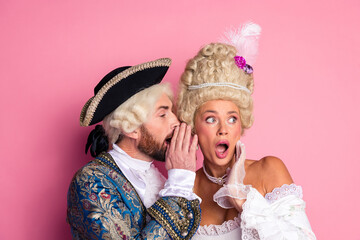 Elegant couple dressed in royal historical costumes sharing a secret against pink backdrop